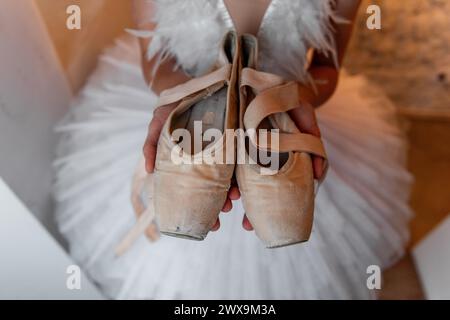 Jeune ballerine en tutu blanc, accessoire de diadème de cheveux à plumes se tient soigneusement, tenant des chaussures de pointe usées, évoquant le sens du dévouement et le quie Banque D'Images