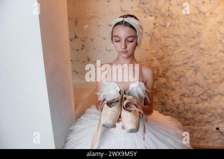 Jeune ballerine en tutu blanc, accessoire de diadème de cheveux à plumes se tient soigneusement, tenant des chaussures de pointe usées, évoquant le sens du dévouement et le quie Banque D'Images