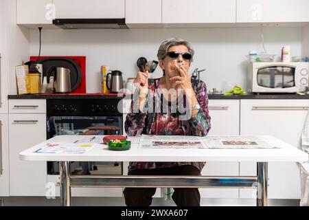 Une vieille dame pleine de rides fume confortablement à la maison dans sa cuisine portant des lunettes de soleil noires. Banque D'Images