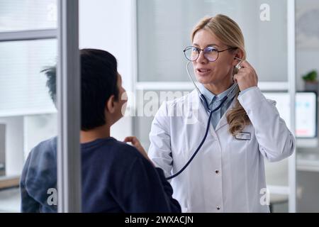 Portrait de taille haute de médecin féminin souriant examinant garçon dans la clinique pédiatrique et en utilisant stéthoscope écoutant l'espace de copie de respiration Banque D'Images