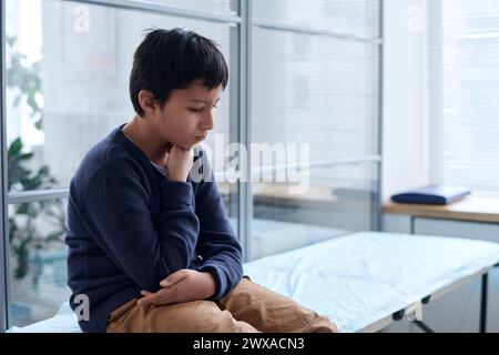 Portrait vue de côté d'un garçon du moyen-Orient en détresse assis sur un lit d'examen dans la clinique et en attente de l'espace de copie du médecin Banque D'Images