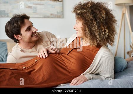 Femme enceinte avec mari allongé dans le lit, homme touchant son ventre de femme et souriant Banque D'Images