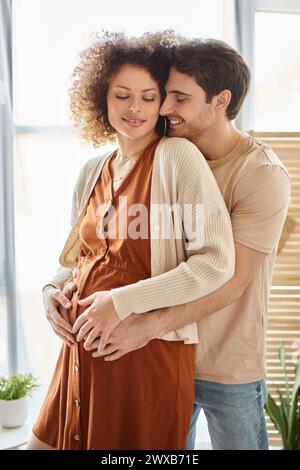 Bel homme heureux embrassant sa femme enceinte de derrière debout à la maison attendant bébé Banque D'Images