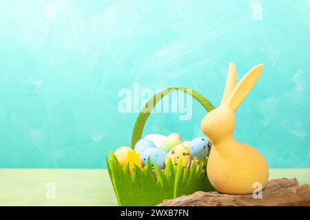 Lapin de Pâques jaune sur vieux bois flotté et panier en feutre vert avec des œufs colorés décoratifs sur fond blanc turquoise. Modèle de carte de voeux de Pâques Banque D'Images