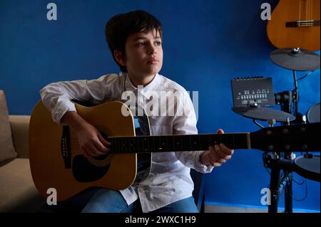 Adolescent sérieux exécutant une mélodie sonore, chantant une chanson tout en jouant de la guitare acoustique dans un studio musical moderne à domicile. Talentueux étalon d'âge élémentaire Banque D'Images