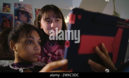 Adolescent asiatique se trouve sur le lit et regarde le contenu avec un ami. Fille afro-américaine assise au sol, surfe Internet en utilisant la tablette. Les filles multiculturelles passent du temps libre ensemble à la maison. Relation amicale. Banque D'Images