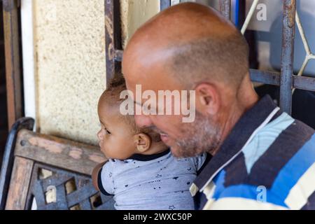 enfant africain et père caucasien assis sur un banc dans la cour au coucher du soleil, bars antivol en arrière-plan Banque D'Images