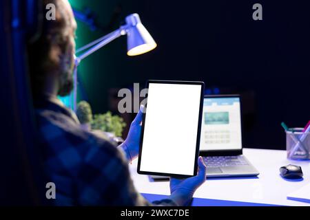 Homme tenant verticalement une tablette avec un écran blanc isolé devant son ordinateur portable. Freelance masculine travaillant à la maison, axée sur l'appareil numérique affichant un modèle de maquette chromakey vierge. Banque D'Images