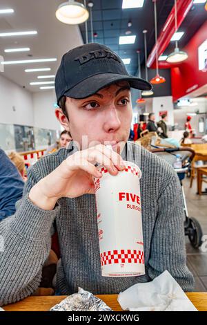 Un adolescent boit une boisson pétillante via une paille dans une tasse en papier jetable dans un restaurant Five Guys Banque D'Images