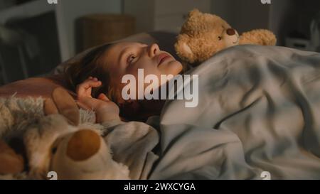 Belle jeune fille se trouve sur le lit sous une couverture avec des jouets en peluche et se repose dans sa chambre le soir. Mignonne écolière se réveille et regarde autour tard dans la nuit. Maison confortable avec intérieur élégant et confortable. Banque D'Images