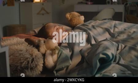 Belle jeune fille se trouve sur le lit sous une couverture avec des jouets en peluche et se repose dans sa chambre le soir. Mignonne écolière se réveille et regarde autour tard dans la nuit. Maison confortable avec intérieur élégant et confortable. Banque D'Images