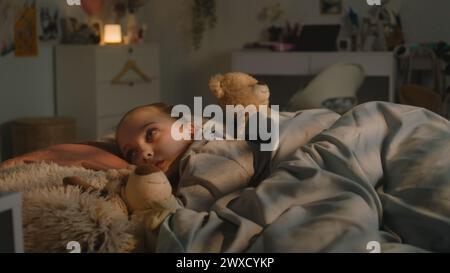 Belle jeune fille se trouve sur le lit sous une couverture avec des jouets en peluche et se repose dans sa chambre le soir. Mignonne écolière se réveille et regarde autour tard dans la nuit. Maison confortable avec intérieur élégant et confortable. Banque D'Images