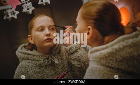 Jeune fille caucasienne fait le maquillage des sourcils debout et regardant dans le miroir dans une chambre confortable et confortable. Adolescente passant du temps libre et s'amusant à la maison la nuit. Concept de style de vie. Banque D'Images