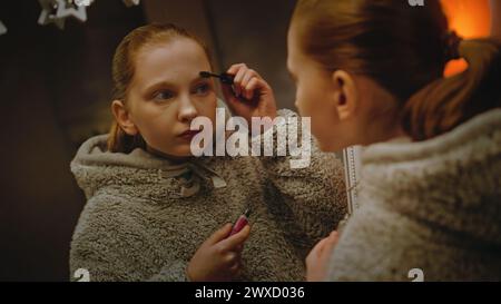 Jeune fille caucasienne fait le maquillage des sourcils debout et regardant dans le miroir dans une chambre confortable et confortable. Adolescente passant du temps libre et s'amusant à la maison la nuit. Concept de style de vie. Banque D'Images