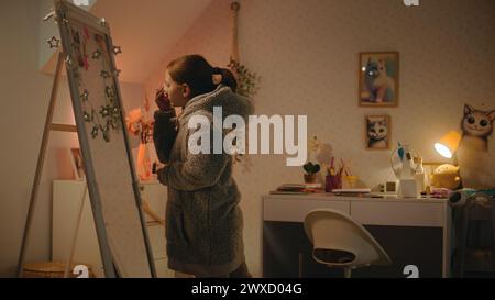 Jeune fille caucasienne fait le maquillage des sourcils debout et regardant dans le miroir dans une chambre confortable et confortable. Adolescente passant du temps libre et s'amusant à la maison la nuit. Concept de style de vie. Banque D'Images