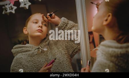 Jeune fille caucasienne fait le maquillage des sourcils debout et regardant dans le miroir dans une chambre confortable et confortable. Adolescente passant du temps libre et s'amusant à la maison la nuit. Concept de style de vie. Banque D'Images