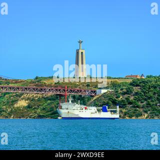 Ferry blanc naviguant sous le pont suspendu en acier 25 de Abril de Lisbonne et le monument religieux et sanctuaire dédié au Sacré-cœur de Jésus Banque D'Images