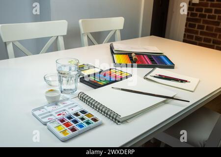 Aquarelles, ustensiles de travail, pinceaux et un cahier pour peindre des aquarelles sur une table blanche à l'intérieur d'une maison Banque D'Images
