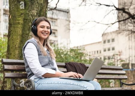 Jeune femme d'affaires réussie assise dans le parc sur le banc travaillant sur un ordinateur portable tout en écoutant podcast sur un casque sans fil. Professionnel féminin Banque D'Images