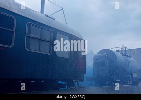 Un train est couvert de pluie et est garé sur une voie ferrée. Le train est entouré d'autres trains et d'un grand pétrolier. La scène est terne et humide, wi Banque D'Images