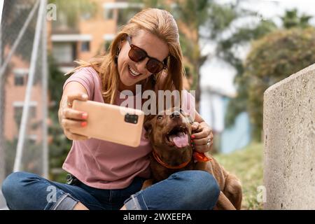 Une femme heureuse portant des lunettes de soleil sourit alors qu'elle prend un selfie avec son chien adorant. Banque D'Images