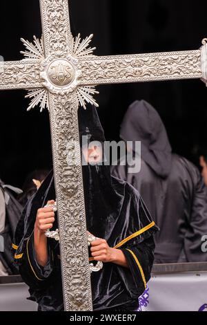 Antigua, Guatemala. 29 mars 2024. Portant le chapeau pointu capirote, un pénitent tient une croix d'argent alors qu'il attend devant l'église Escuela de Cristo le début de la procession du vendredi Saint Senor Sepultado pendant Semana Santa, le 29 mars 2024 à Antigua, Guatemala. La procession opulente est l'une des plus grandes au monde impliquant des milliers de dévots et dure 12 heures. Crédit : Richard Ellis/Richard Ellis/Alamy Live News Banque D'Images