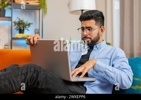 Bouleversé jeune homme indien assis sur le canapé à la maison à l'aide d'un ordinateur portable ayant l'anxiété et le stress. Un homme hispanique barbu choqué réagissant à la perte, de mauvaises nouvelles sur netbook dans l'appartement du salon. Virus informatique. Banque D'Images