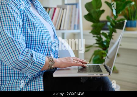Gros plan des mains de la femme mature tapant sur ordinateur portable, fond intérieur de la maison Banque D'Images