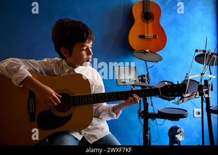 Adolescent jouant de la guitare dans le studio de musique moderne. Guitare électrique accrochée à un mur bleu près de la batterie. Les gens. Leçon de musique. Éducation des enfants et Banque D'Images