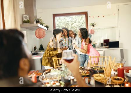 Jeune groupe joyeux d'amis divers tenant des boissons et serrant dans les bras à la cuisine à la maison pendant le temps de la fête Banque D'Images
