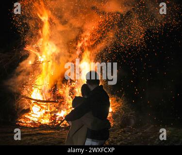 Sythen, NRW, Allemagne. 30 mars 2024. Un câlin de couple, debout près du feu de joie annuel de Pâques à la ferme Prickingshof à Sythen, Westfalia (Münsterland). L'allumage des feux de joie de Pâques est une tradition depuis de nombreux siècles en Allemagne, généralement le samedi Saint ou le dimanche de Pâques. Les services d’incendie et d’urgence veillent à ce que le feu soit protégé pendant que les bénévoles ont soigneusement inspecté le bois de chauffage pour s’assurer qu’aucune faune ne s’est réfugiée dans les branches. Crédit : Imageplotter/Alamy Live News Banque D'Images