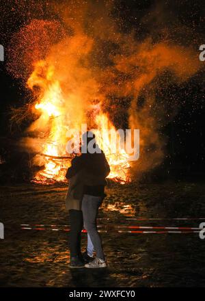 Sythen, NRW, Allemagne. 30 mars 2024. Un câlin de couple, debout près du feu de joie annuel de Pâques à la ferme Prickingshof à Sythen, Westfalia (Münsterland). L'allumage des feux de joie de Pâques est une tradition depuis de nombreux siècles en Allemagne, généralement le samedi Saint ou le dimanche de Pâques. Les services d’incendie et d’urgence veillent à ce que le feu soit protégé pendant que les bénévoles ont soigneusement inspecté le bois de chauffage pour s’assurer qu’aucune faune ne s’est réfugiée dans les branches. Crédit : Imageplotter/Alamy Live News Banque D'Images