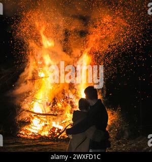 Sythen, NRW, Allemagne. 30 mars 2024. Un câlin de couple, debout près du feu de joie annuel de Pâques à la ferme Prickingshof à Sythen, Westfalia (Münsterland). L'allumage des feux de joie de Pâques est une tradition depuis de nombreux siècles en Allemagne, généralement le samedi Saint ou le dimanche de Pâques. Les services d’incendie et d’urgence veillent à ce que le feu soit protégé pendant que les bénévoles ont soigneusement inspecté le bois de chauffage pour s’assurer qu’aucune faune ne s’est réfugiée dans les branches. Crédit : Imageplotter/Alamy Live News Banque D'Images