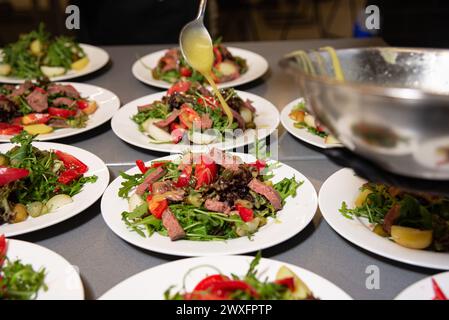 Le chef vinaigrette avec précision sur des salades de steak, superbement disposées sur des assiettes dans une cuisine commerciale animée. Banque D'Images