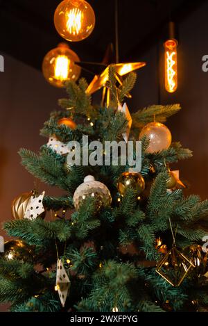 Sapin de Noël festif orné de décorations dorées et blanches, agrémenté d'élégantes ampoules Edison. Banque D'Images