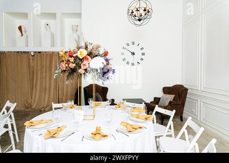 Table sophistiquée pour un événement avec somptueuse pièce maîtresse fleurie, chaises blanches et horloge murale moderne. Banque D'Images