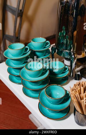 Des piles de tasses à café et de soucoupes turquoises élégantes sont installées à côté d'une urne à café brillante, prête pour le service. Banque D'Images