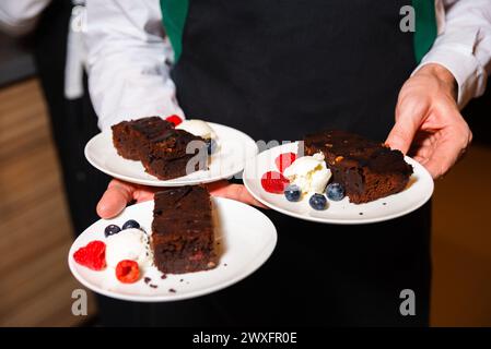 Un serveur présente des assiettes de riches brownies au chocolat accompagnées de baies fraîches et d'une cuillère de crème, prêtes pour le service de dessert. Banque D'Images