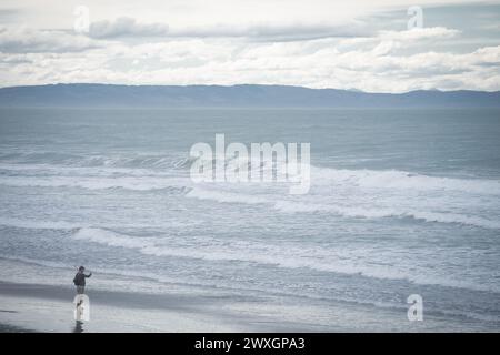 Homme seul prenant des photos sur la plage alors que de grosses vagues approchent, Nouvelle-Zélande. Banque D'Images