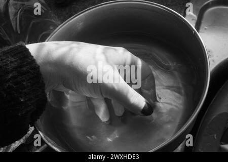 Laver une main - tir noir et blanc d'une main à l'intérieur d'une casserole en métal immergée dans l'eau avec une lumière et une ombre contrastées. Banque D'Images