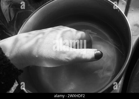 Laver une main - tir noir et blanc d'une main à l'intérieur d'une casserole en métal immergée dans l'eau avec une lumière et une ombre contrastées. Banque D'Images