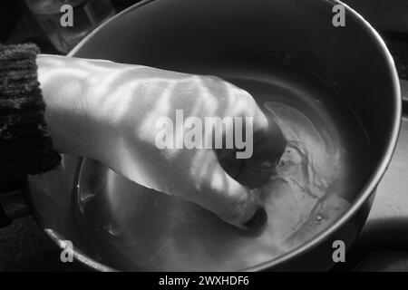 Laver une main - tir noir et blanc d'une main à l'intérieur d'une casserole en métal immergée dans l'eau avec une lumière et une ombre contrastées. Banque D'Images