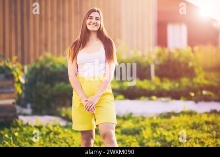 Les bermudas jaunes sont portés par la jeune femme brune caucasienne qui pose pour la photographie en souriant tout en se tenant dans la cour arrière de la maison de campagne à la fin Banque D'Images