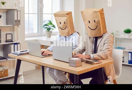 Des gens d'affaires drôles sans visage dans des sacs en papier sur la tête avec des visages souriants dessinés travaillant sur des ordinateurs portables. Banque D'Images