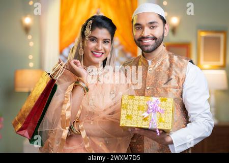 Heureux couple musulman indien souriant tenant des sacs à provisions et une boîte cadeau en main en regardant la caméra à la maison - concept de promotion du festival, Ramadan Banque D'Images