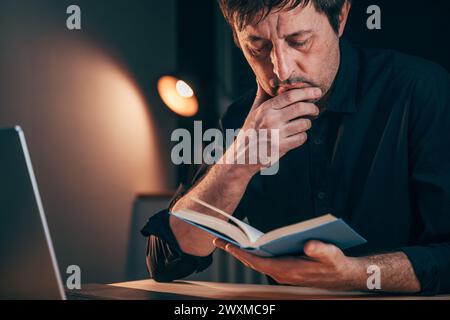 Homme d'affaires lisant un livre tout en étant assis au bureau tard dans la nuit, focus sélectif Banque D'Images