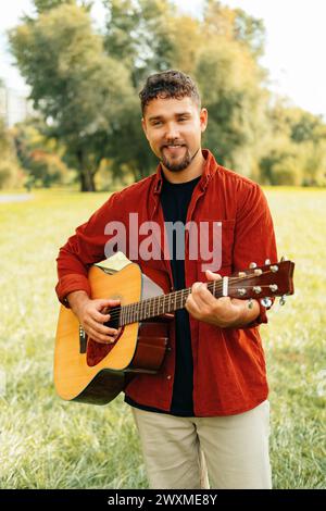 Plan vertical extérieur d'un bel homme tenant et jouant d'une guitare acoustique. Banque D'Images