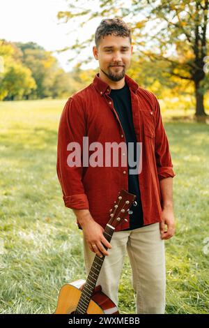 Plan vertical extérieur d'un bel homme tenant une guitare acoustique dans le parc tout en marchant. Banque D'Images