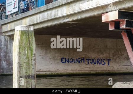 Graffiti disant « ASSEZ DE TOURISTES » sous un pont de canal avec des vélos stationnés au-dessus, montrant le sentiment d'Amsterdam fatigué du tourisme dans le centre-ville Banque D'Images