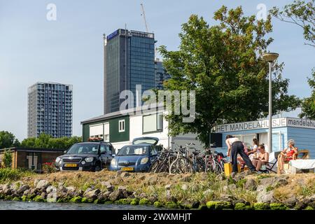 Les gens se détendent au bord de l'eau près des vélos et des voitures garés à Amsterdam Noord, avec des immeubles modernes de grande hauteur en arrière-plan par une journée ensoleillée Banque D'Images